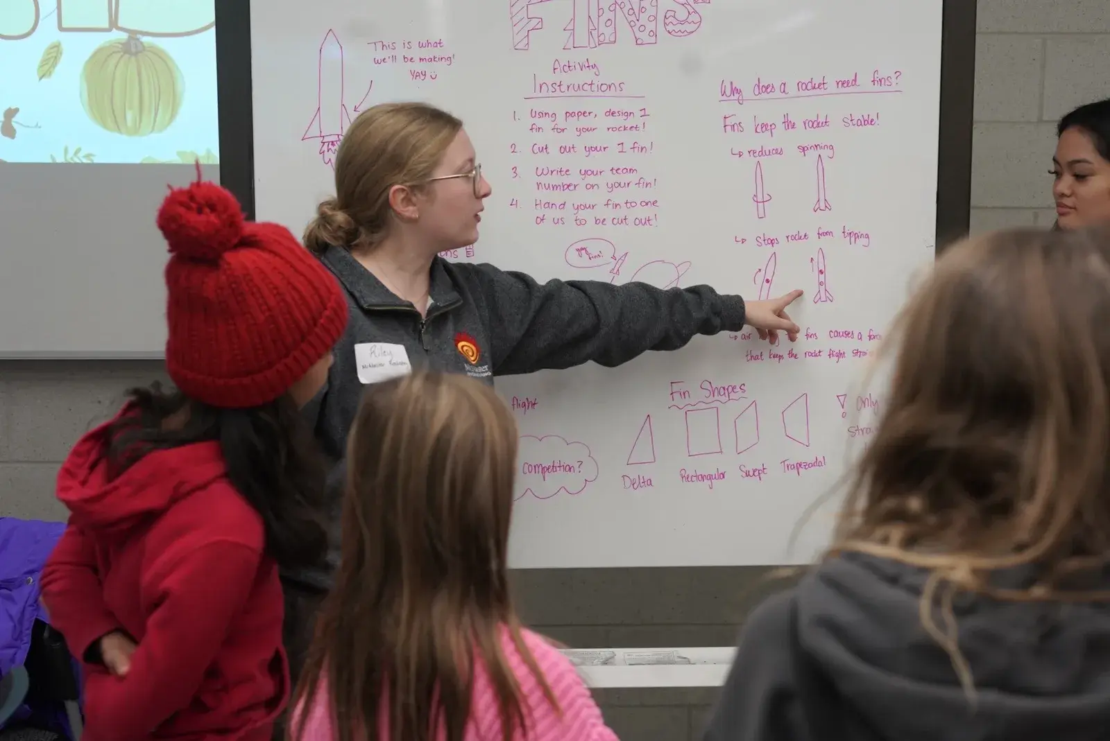 A team member at a whiteboard labelled FINS explains rocket stability to a group of children during a CAGIS STEM session.