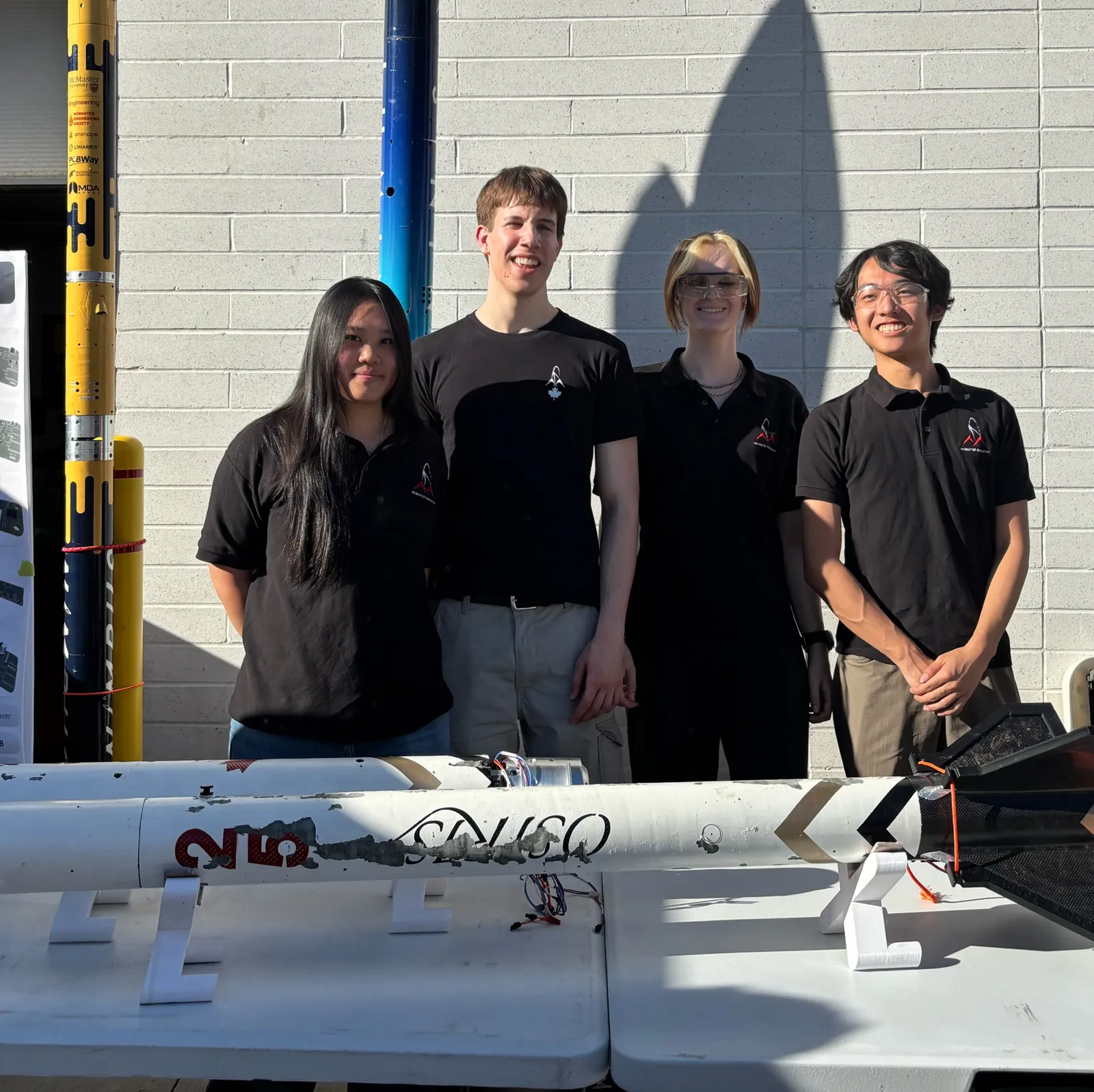Four team members in black McMaster Rocketry shirts standing behind the Osiris airframe on stands at the Clubsfest 2026 table, with two rockets upright to the left.