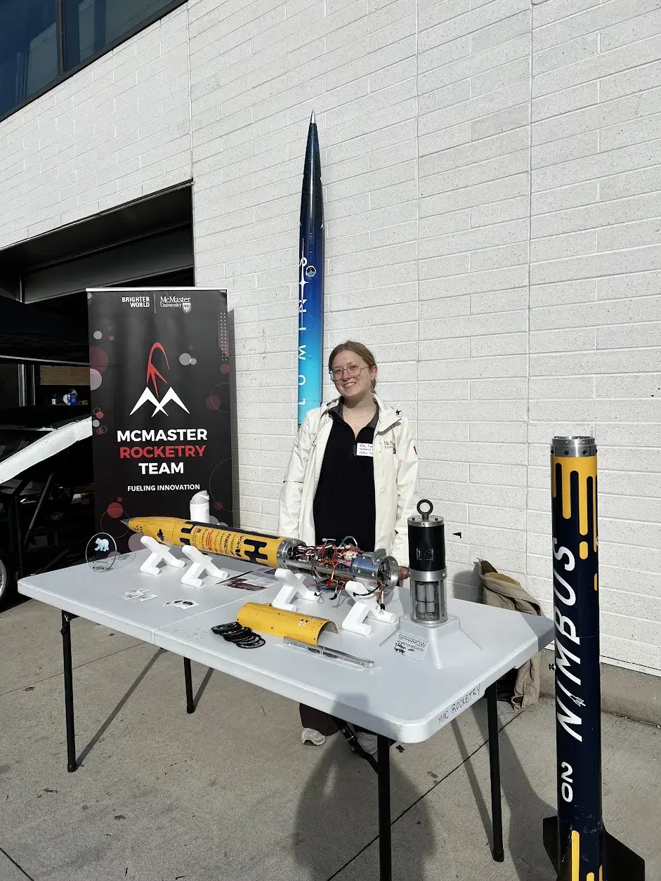 A team member at the outdoor Clubsfest table with the Osiris airframe open on stands, a rocket against the wall, and Nimbus standing beside the booth.