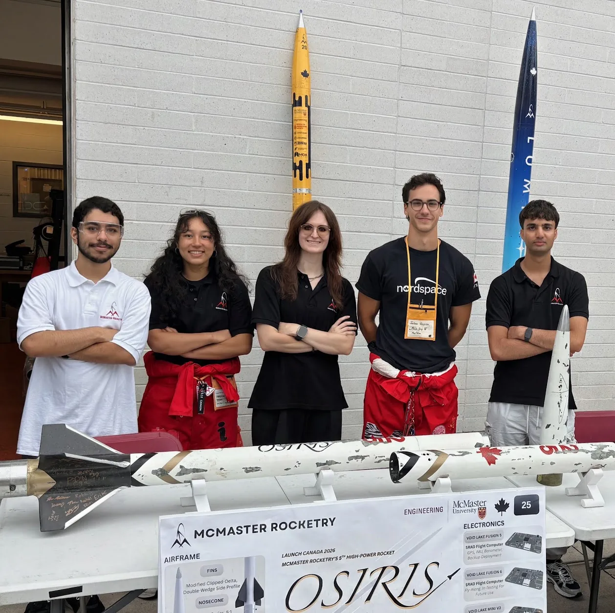 Five team members beside the Osiris and Void Lake airframes and two standing rockets at the Facultyfest 2026 table.