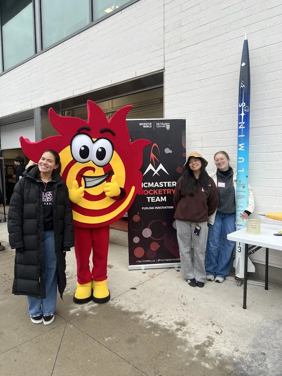 Three team members and the McMaster mascot beside the team banner and a standing Luminis airframe at Facultyfest.