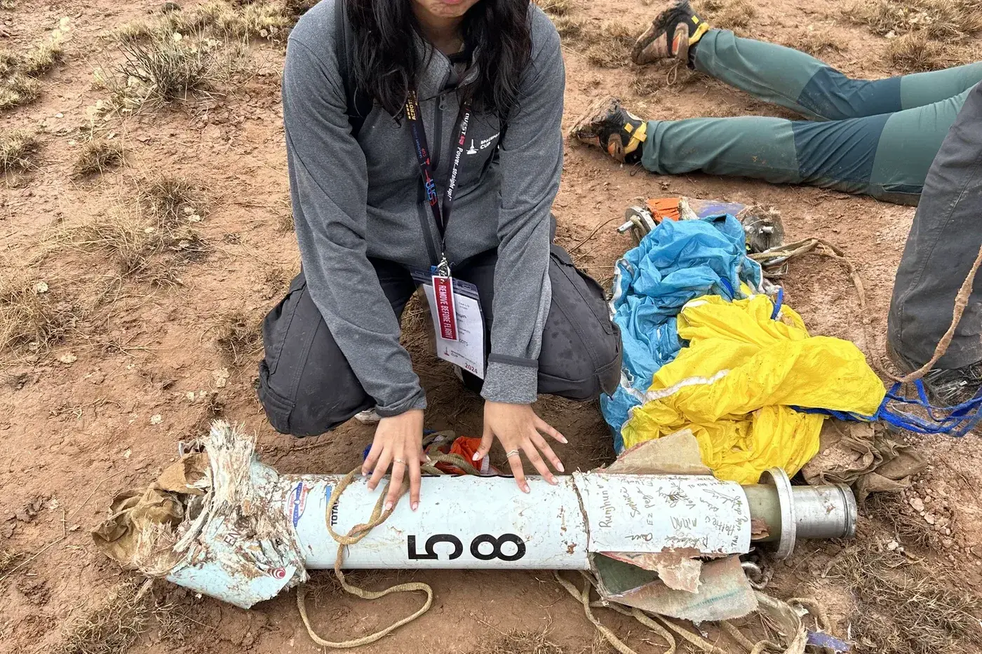 A team member kneels beside the recovered Luminis airframe after landing, bib 58 and signed fins visible beside the parachute.