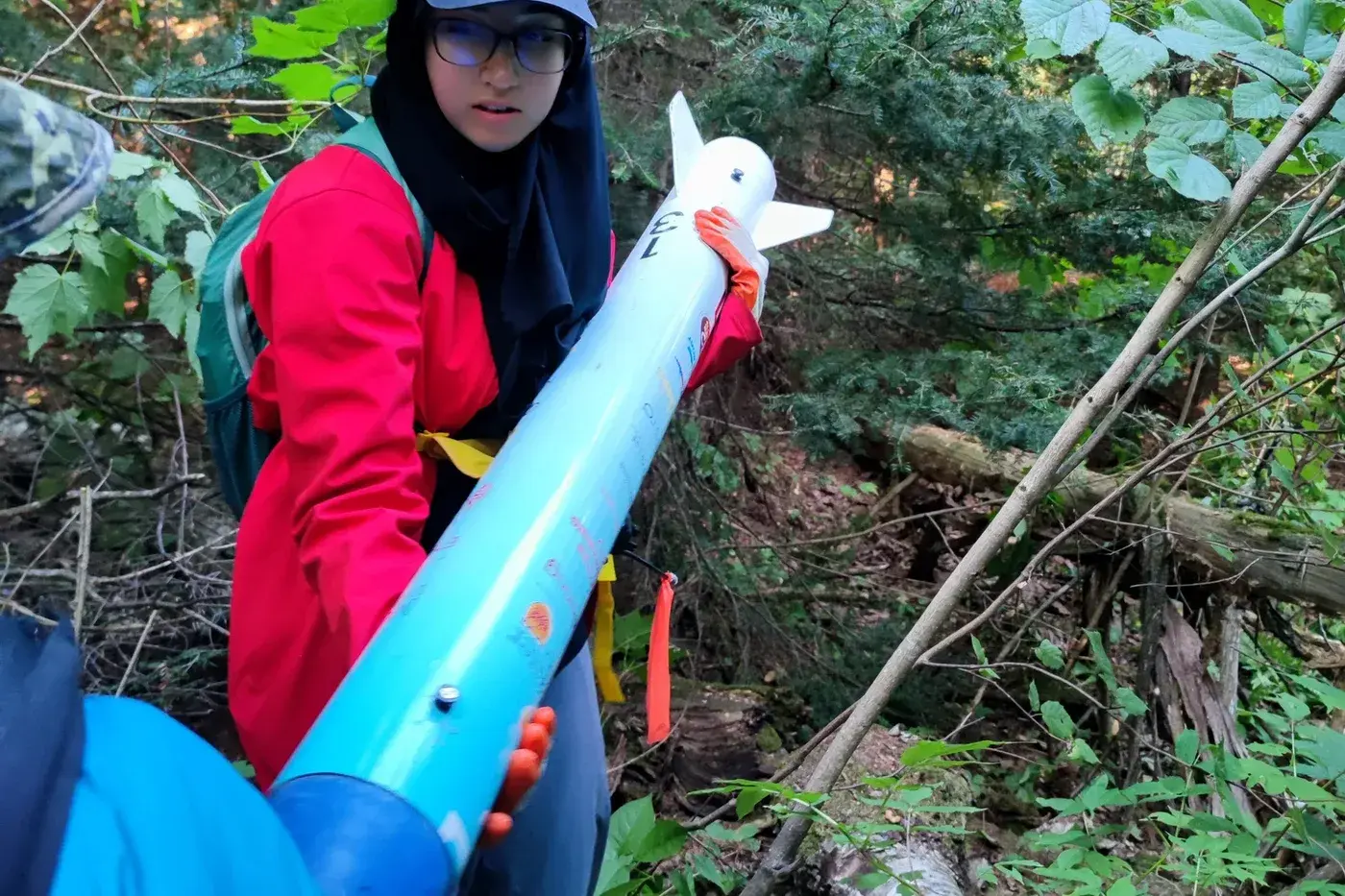 A team member carries the recovered Luminis V2 airframe through the bush, bib 13 on the tail and orange streamer on the tube.