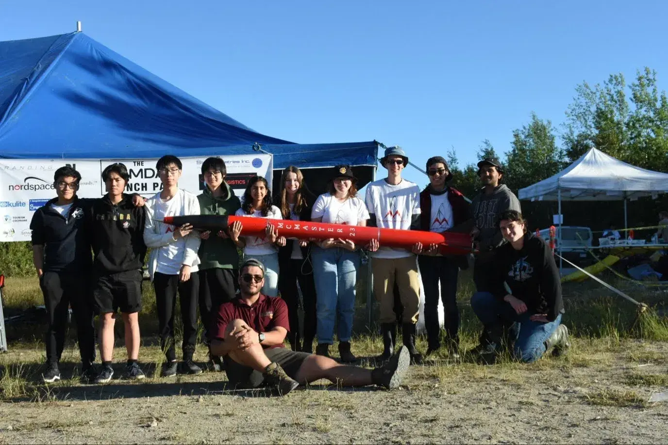 The Marauder I team stands with the completed red airframe under the Launch Canada tent, McMaster 23 lettering visible.