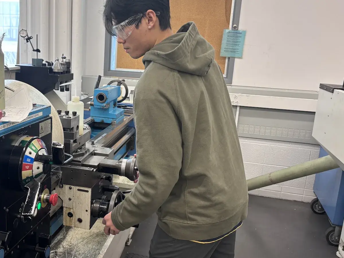 A team member at the lathe, one hand on the Colchester carriage handwheel and safety glasses on, with the tool post and swarf on the bed in front of him.