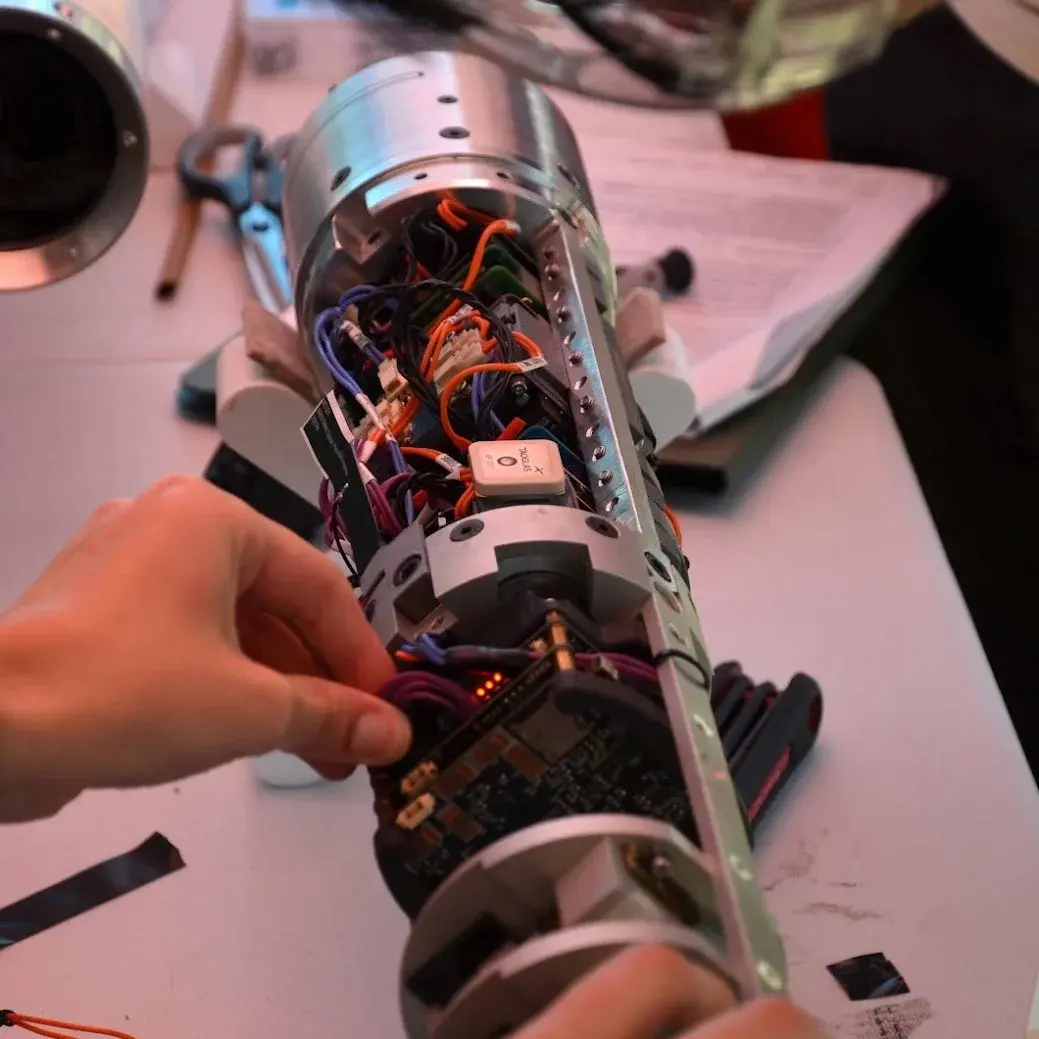 Hands wiring an open avionics bay on the workbench: coloured harnesses, a circuit board with red indicator LEDs, and tools laid out beside it.