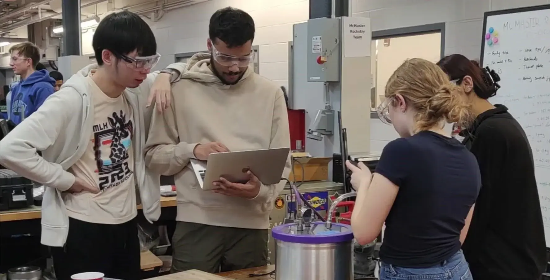 Team members around a vacuum chamber in the composites bay: one films the setup on a phone while others monitor the laptop and gauge.